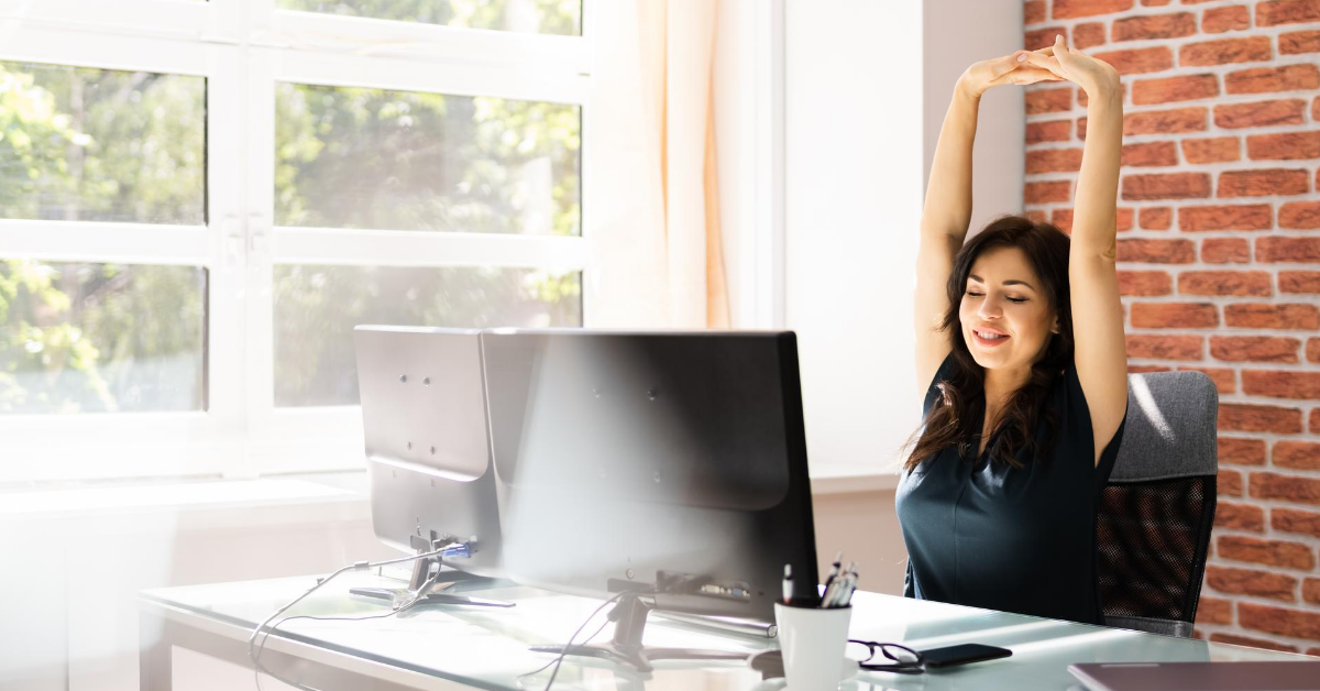 Woman in an office setting, sitting behind a desk, stretching her arms.