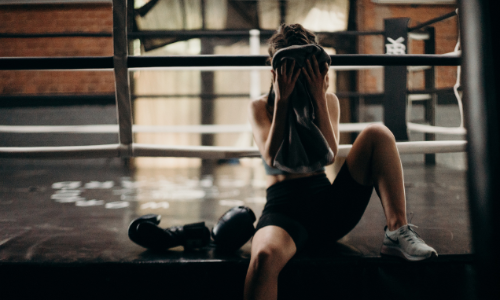 Woman sitting down by boxing ring, wiping the sweat on her face with a towel.