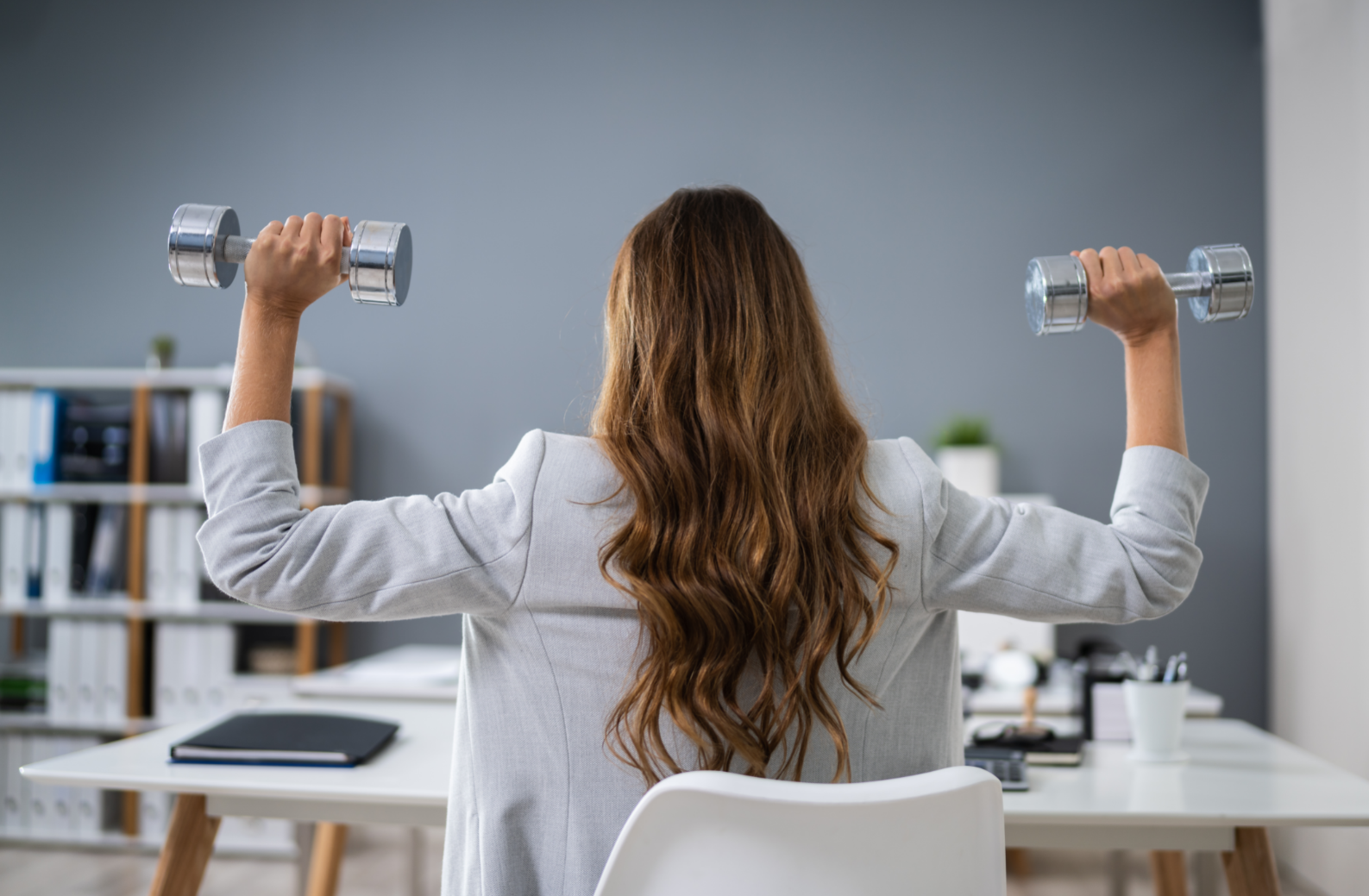 Woman sitting at a desk, with her back turned to the camera, lifting two small weights.