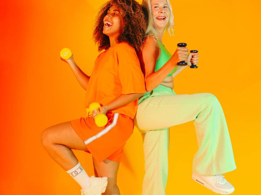 Younger and older woman wearing bright orange and lime green athlethic clothes, smiling while holding dumbbells.