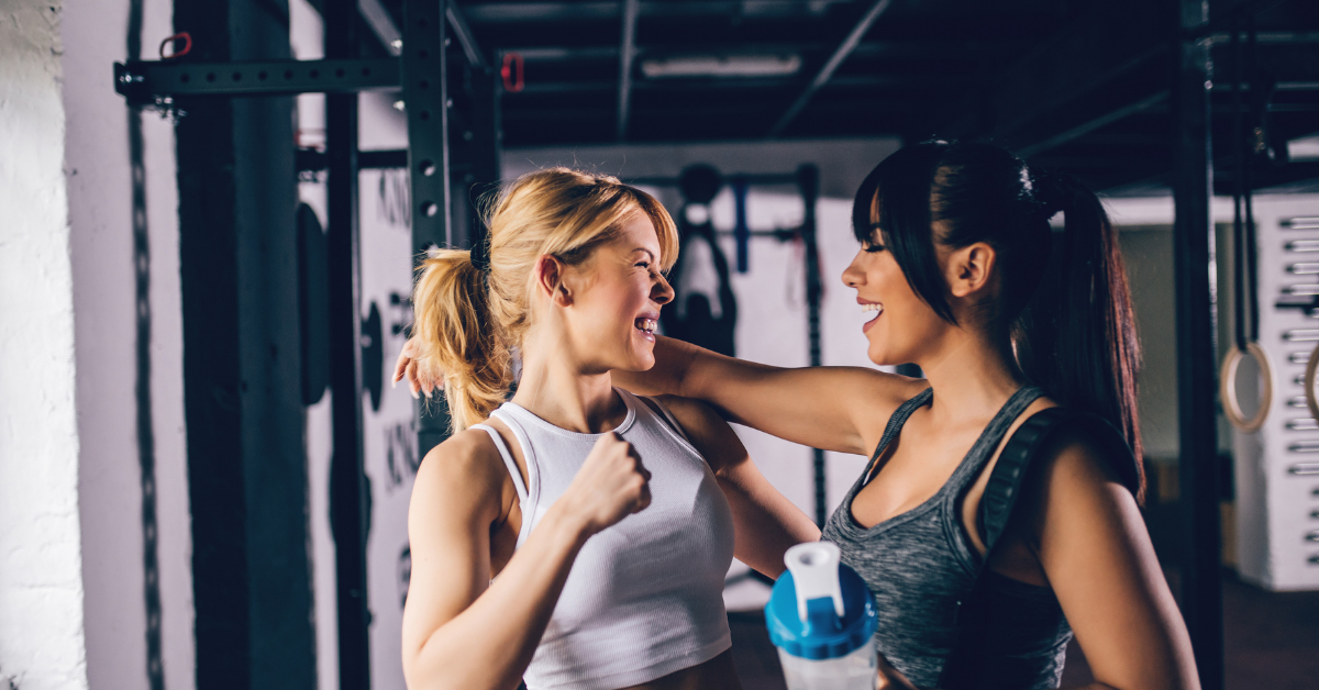 Two women at the gym wearing athletic clothes and talking to each other.
