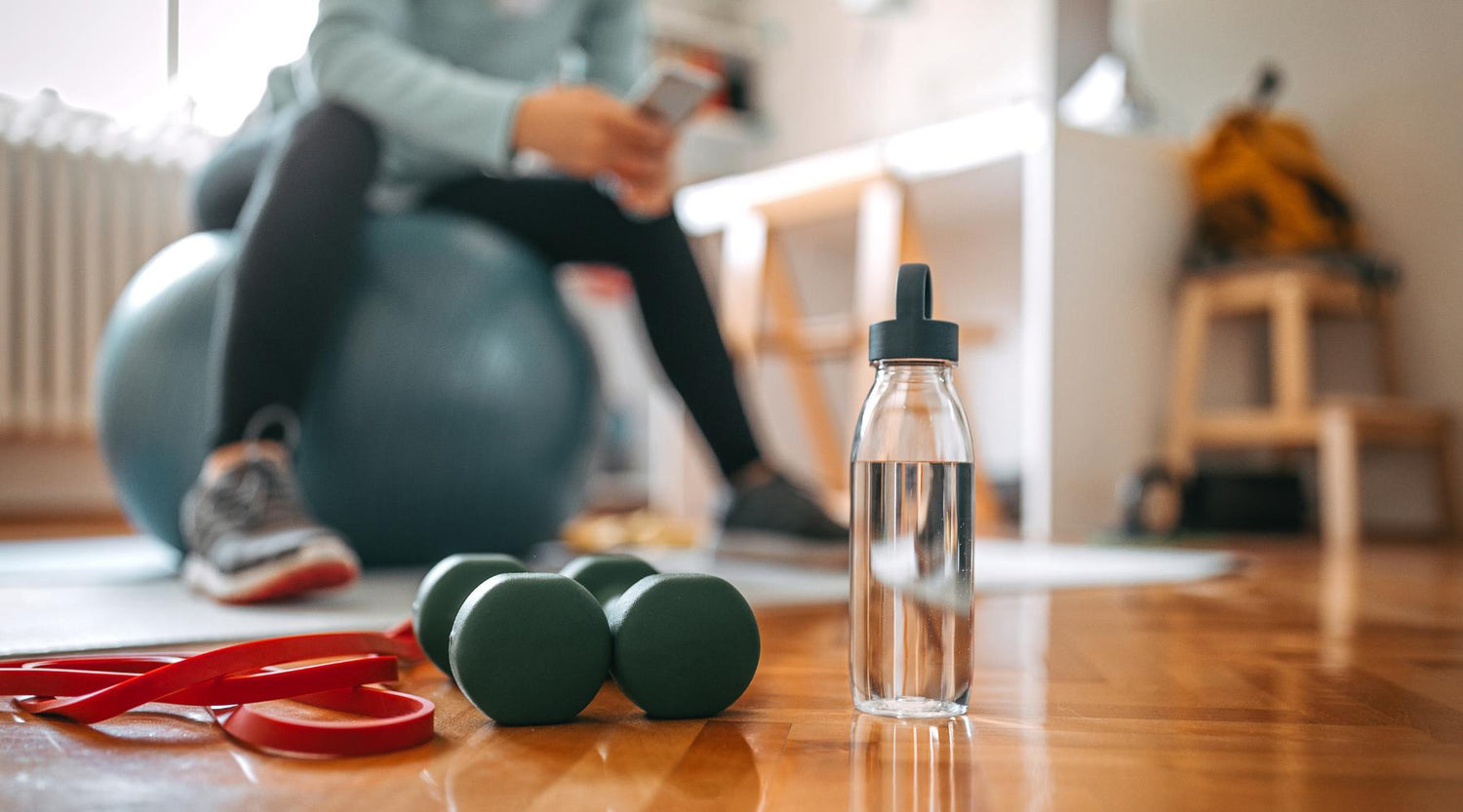 Woman browsing her phone while sitting on a yoga ball in her living room with a red resistance band and water bottle in the foreground