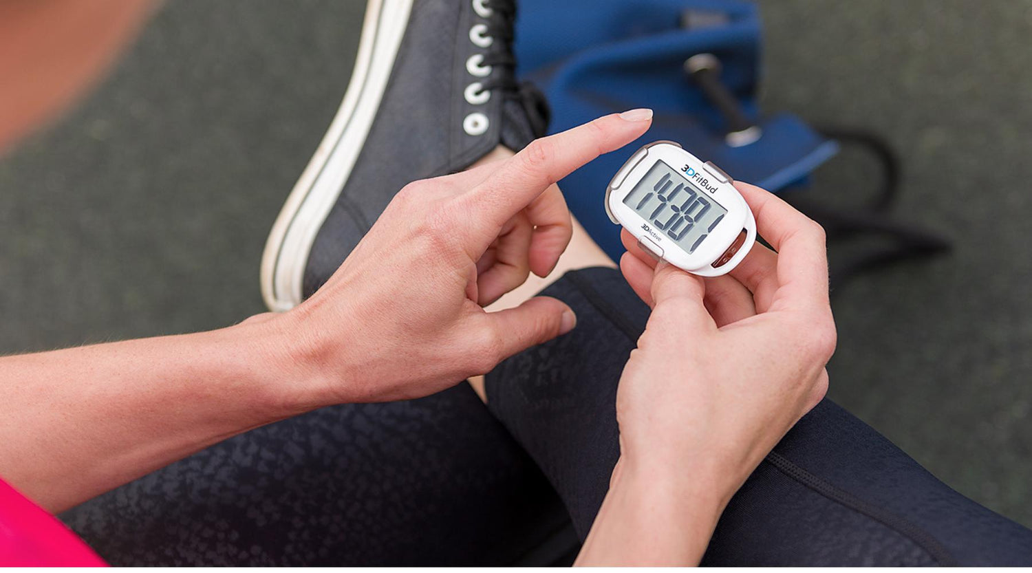 A person sitting outdoors checking their step count on a 3DActive digital pedometer while wearing black athletic shoes and leggings.