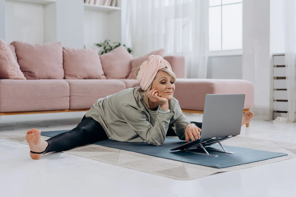 Older woman in yoga clothes stretching legs on living room floor while watching something on a laptop.