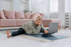 Older woman in yoga clothes stretching legs on living room floor while watching something on a laptop.