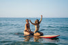 Two women sitting on a surf board in the ocean.