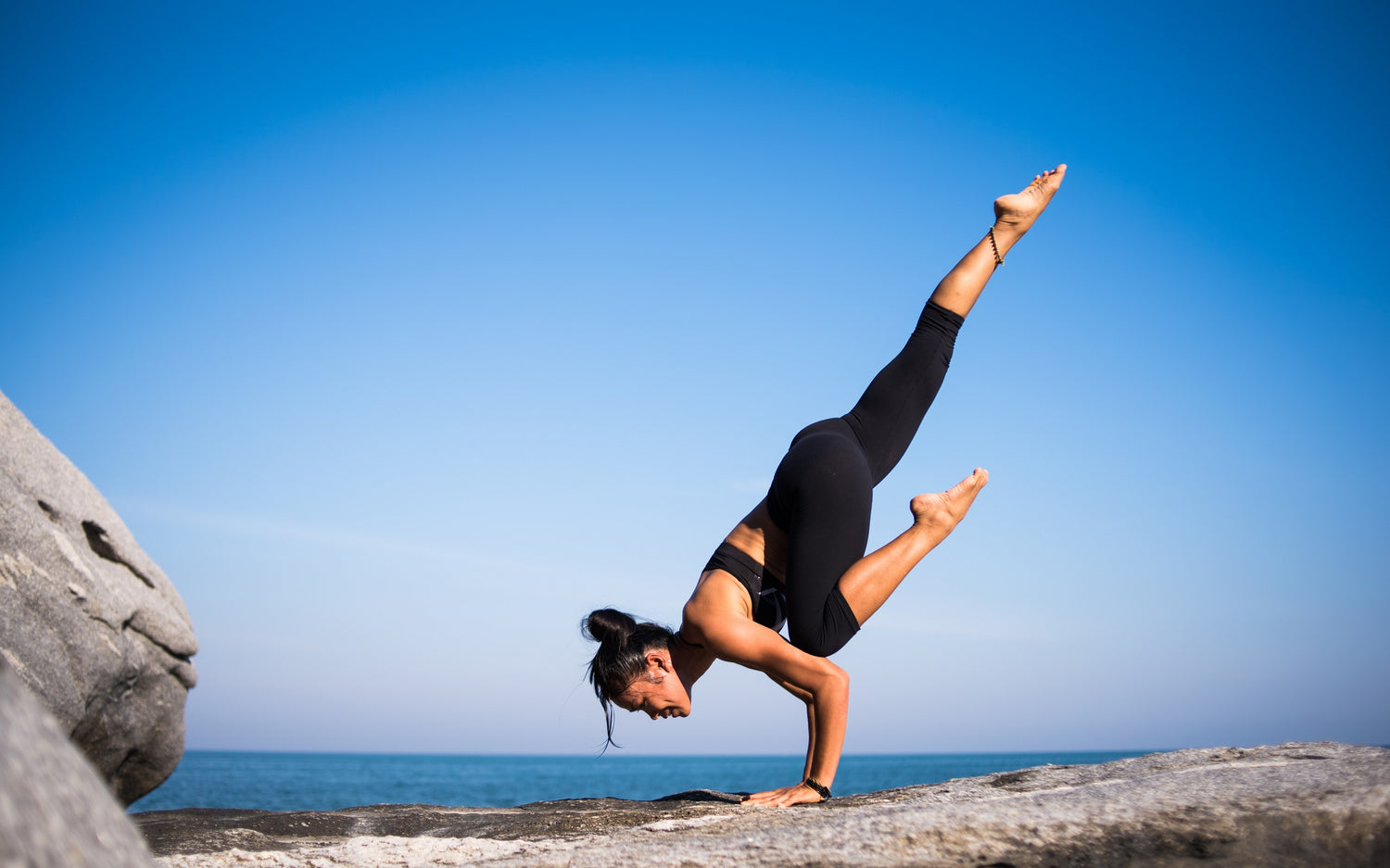 Woman doing a yoga pose on a rocky surface with a clear blue sky behind her.